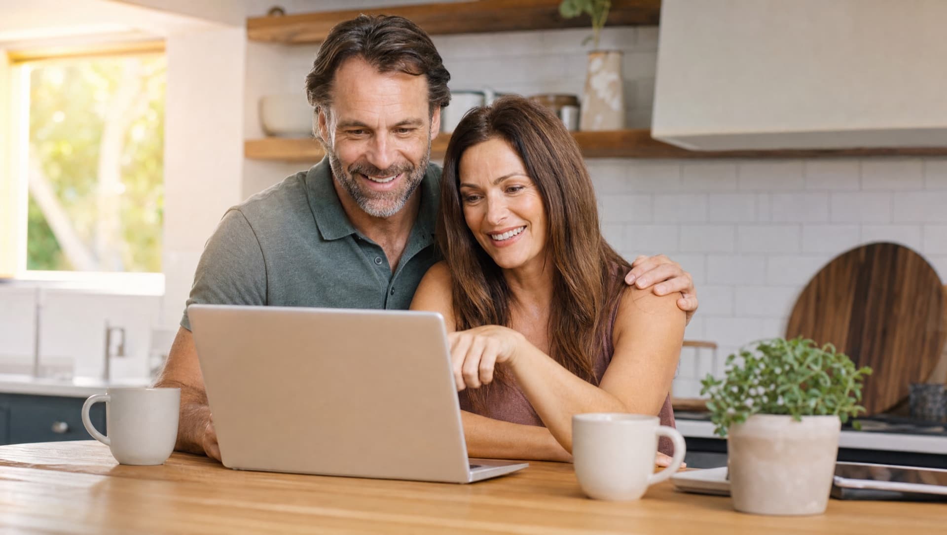 A couple reviewing plans on a laptop in a sunlit kitchen, both smiling